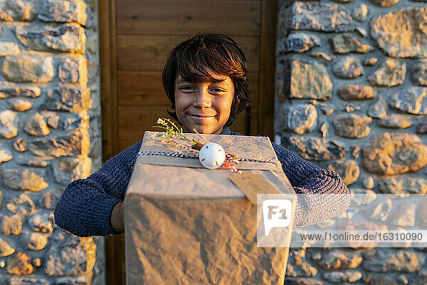 Nahaufnahme eines lächelnden Jungen  der ein Weihnachtsgeschenk vor einem Haus bei Sonnenuntergang hält