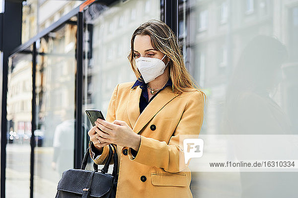 Frau mit Schutzmaske benutzt ihr Smartphone  während sie auf öffentliche Verkehrsmittel wartet