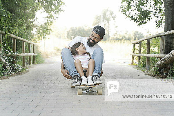 Lächelnder Vater und Tochter beim Skateboardfahren auf dem Fußweg im Park