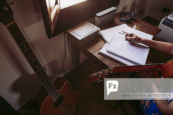 Junge Frau schreibt in ein Buch  während sie im Wohnzimmer Gitarre übt