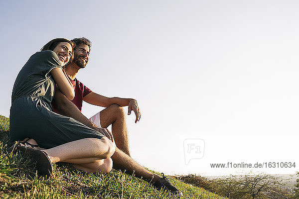 Frau umarmt Mann im Gras sitzend gegen klaren Himmel