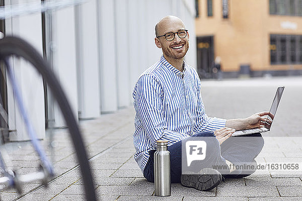 Glücklicher mittlerer Erwachsener  der an einem Laptop arbeitet  während er auf der Straße sitzt
