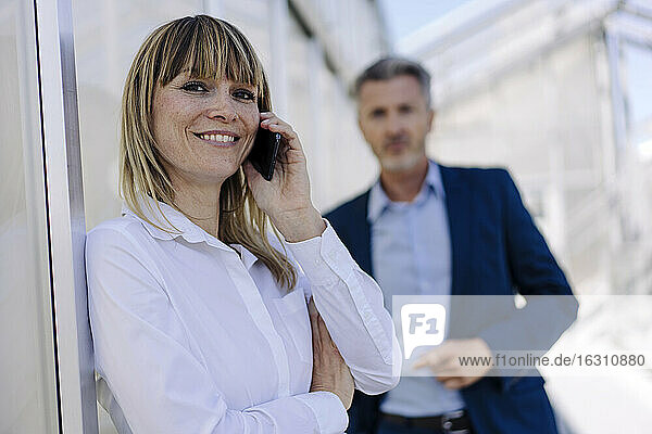 Lächelnde Geschäftsfrau im Gespräch über ein Smartphone  während ein männlicher Mitarbeiter im Hintergrund steht