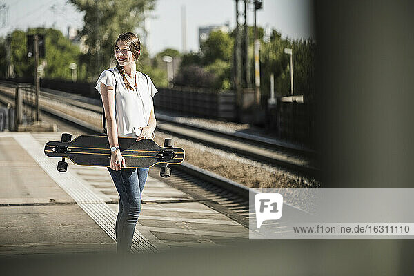 Lächelnde Frau mit Skateboard wartet am Bahnsteig an einem sonnigen Tag