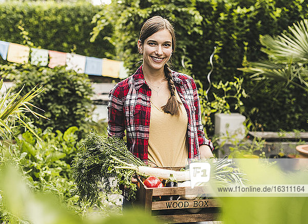 Lächelnde Frau  die Gemüse in einer Kiste trägt  während sie an Pflanzen im Garten steht