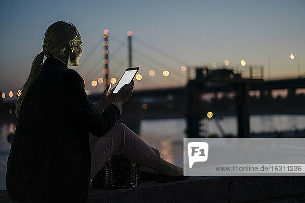 Weibliche Fachkraft  die ein digitales Tablet benutzt  während sie in der Abenddämmerung auf einer Stützmauer sitzt