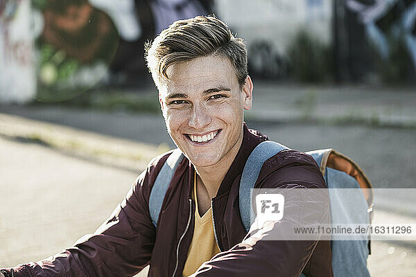 Lächelnder junger Mann in Freizeitkleidung auf der Straße sitzend
