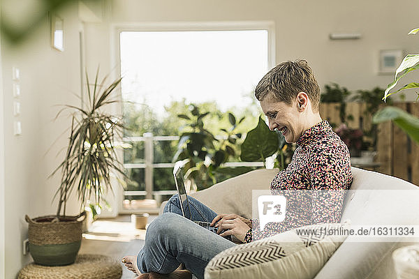 Lächelnde Frau  die einen Laptop benutzt  während sie zu Hause auf dem Sofa im Wohnzimmer sitzt