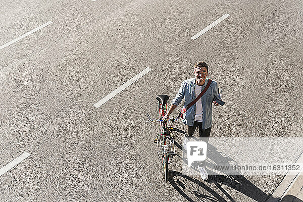 Lächelnder Mann mit Fahrrad beim Überqueren der Straße in der Stadt an einem sonnigen Tag