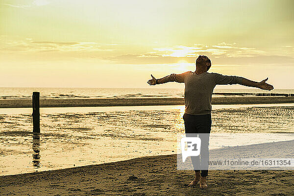 Carefree man with arms outstretched standing at beach against sky during sunset