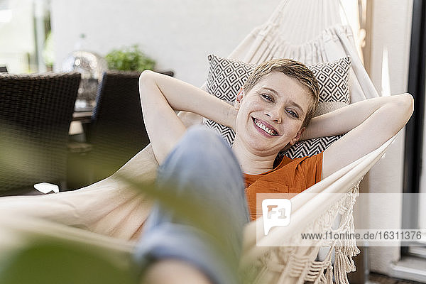 Lächelnde erwachsene Frau mit den Händen hinter dem Kopf auf einer Hängematte auf einer Veranda liegend