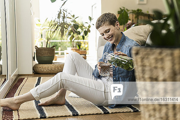 Lächelnde Frau mit kurzen Haaren gießt ein Getränk in ein Glas  während sie zu Hause auf einem Teppich sitzt