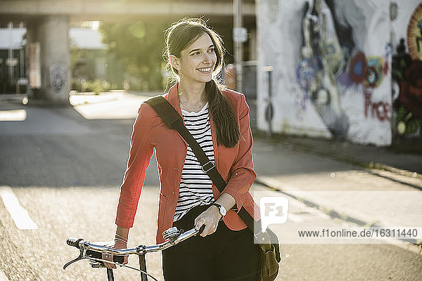 Smiling young woman looking away while walking with bicycle on street in city