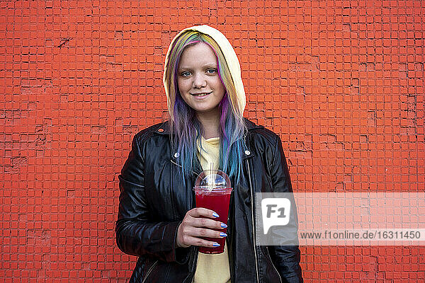 Porträt einer jungen Frau mit gefärbtem Haar und einem Getränk zum Mitnehmen vor einer roten Wand