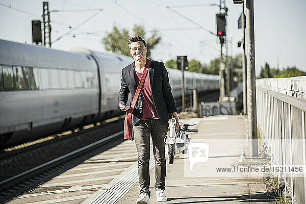 Lächelnder junger Mann mit Tretroller auf dem Bahnsteig