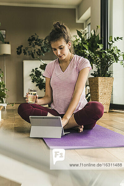 Junge Frau hält Wasser und benutzt ein digitales Tablet  während sie zu Hause auf einer Übungsmatte sitzt