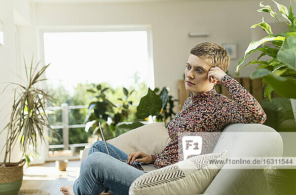 Nachdenkliche Frau im mittleren Erwachsenenalter mit Laptop auf dem Sofa im Wohnzimmer sitzend