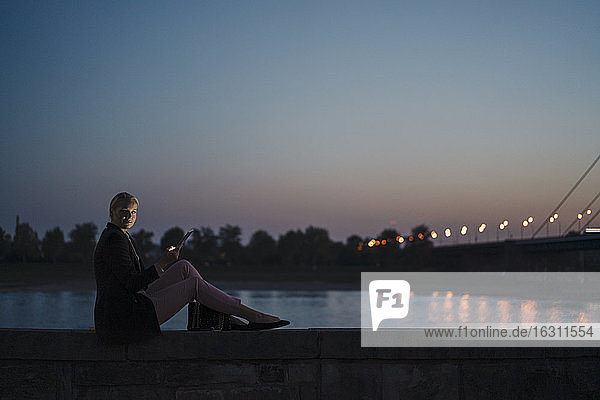 Geschäftsfrau sitzt auf Stützmauer am Fluss gegen klaren Himmel in der Abenddämmerung