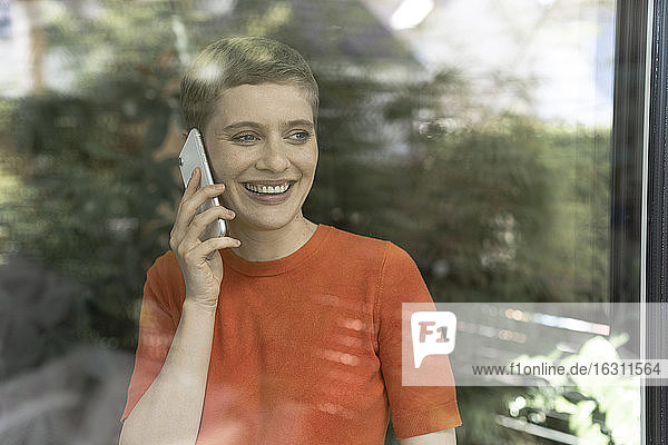 Lächelnde Frau  die über ein Smartphone spricht  während sie zu Hause steht  gesehen durch ein Fenster