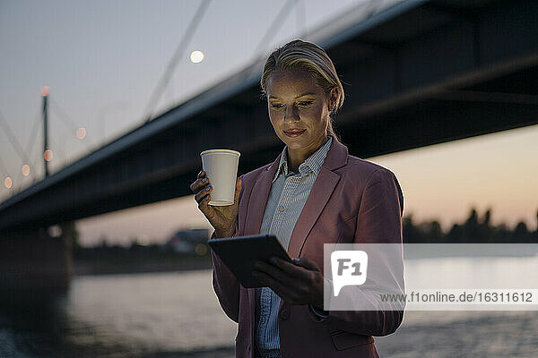 Schöne Geschäftsfrau  die ein digitales Tablet benutzt  während sie einen Einwegbecher vor einer Brücke in der Stadt in der Abenddämmerung hält