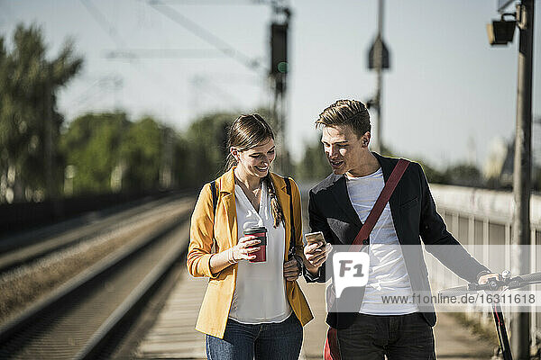 Young man showing smart phone to female friend while walking on railroad station platform