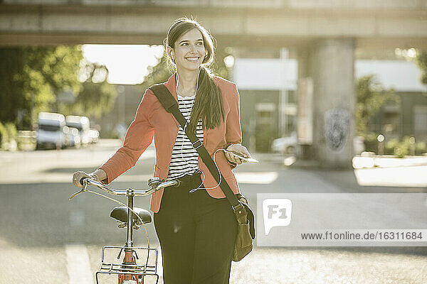 Lächelnde Frau mit Fahrrad hört Musik über ihr Smartphone  während sie auf der Straße geht
