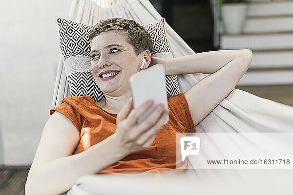Nahaufnahme einer lächelnden schönen Frau mit drahtlosen Kopfhörern und Smartphone  die sich in einer Hängematte entspannt