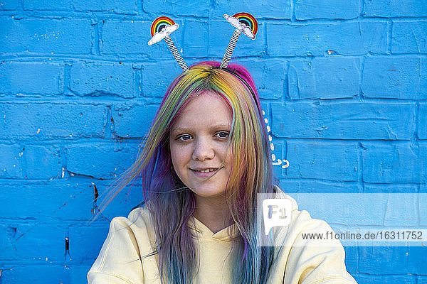 Junge Frau mit gefärbtem Haar und Regenbogen-Alice-Band vor einer blauen Wand