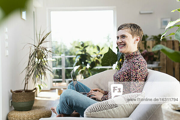 Lächelnde Frau mit Laptop  die wegschaut  während sie auf dem Sofa im Wohnzimmer sitzt