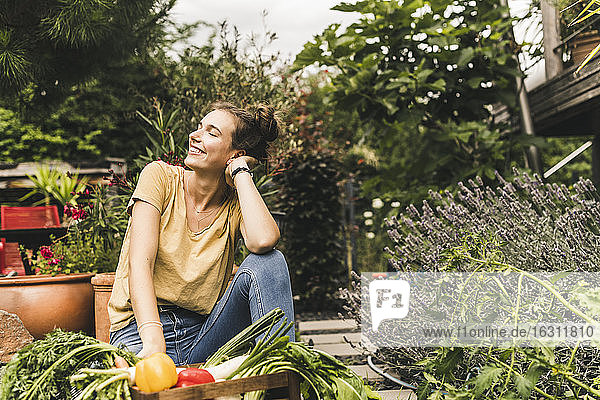 Junge Frau mit geschlossenen Augen sitzt bei Gemüse und Pflanzen im Gemeinschaftsgarten