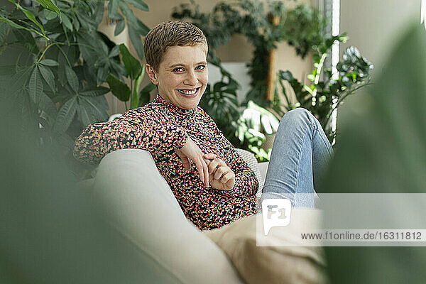 Lächelnde schöne Frau mit kurzen Haaren sitzt auf Sofa gegen Zimmerpflanzen im Wohnzimmer