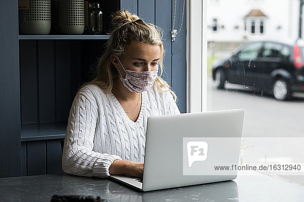 Frau mit Gesichtsmaske  die allein an einem Café-Tisch mit einem Laptop-Computer sitzt und aus der Ferne arbeitet.