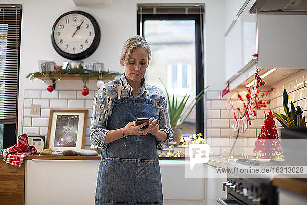 Blonde Frau mit blauer Schürze steht in der Küche und benutzt ein Mobiltelefon.