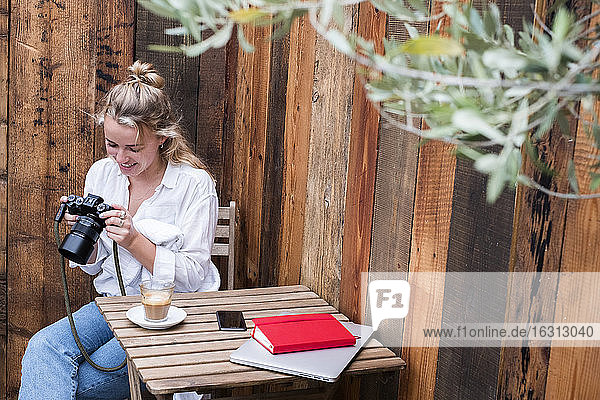 Junge blonde Frau  die allein an einem Café-Tisch sitzt und auf die Anzeige einer Digitalkamera schaut.