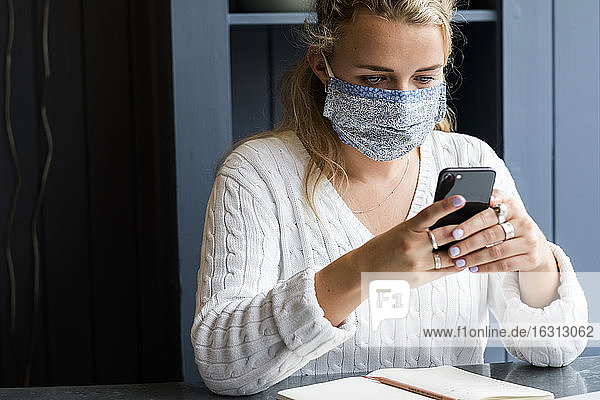 Young blond woman wearing face mask sitting alone in a cafe  using mobile phone  working remotely.