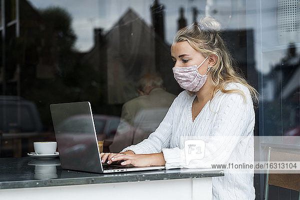 Woman wearing face mask sitting alone at a cafe table with a laptop computer  working remotely.