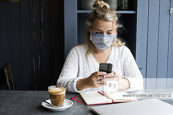 Frau mit Gesichtsmaske sitzt allein an einem Café-Tisch mit einem Laptop-Computer und benutzt ein Mobiltelefon