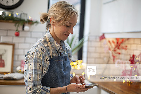 Blonde Frau mit blauer Schürze steht in der Küche und benutzt ein Mobiltelefon.