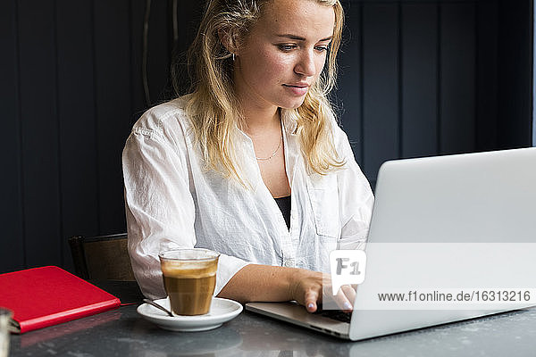 Junge blonde Frau mit Gesichtsmaske sitzt allein mit einem Laptop-Computer an einem Café-Tisch