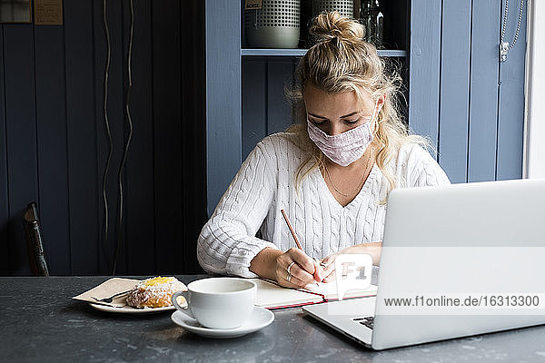 Frau mit Gesichtsmaske sitzt allein mit einem Laptop  schreibt in ein Notizbuch  arbeitet aus der Ferne.