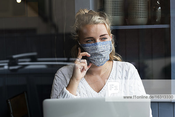 Blond woman wearing face mask sitting alone with a laptop  using mobile phone  working remotely.