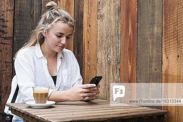 Junge blonde Frau  die allein an einem Café-Tisch im Freien sitzt und ihr Mobiltelefon überprüft.