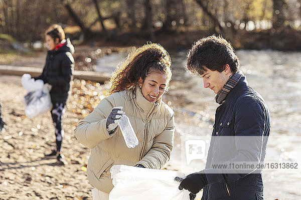 Lächelnde Freiwillige mit männlichem Freund  der eine Plastikflasche im Müllsack hält