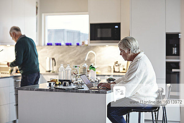 Senior woman using laptop while male preparing food in kitchen