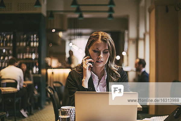 Businesswoman talking on phone while looking at laptop in restaurant