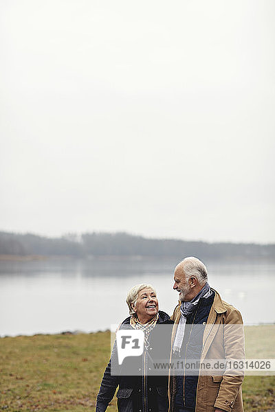 Lächelndes älteres Paar schaut sich an  während es am See vor klarem Himmel steht