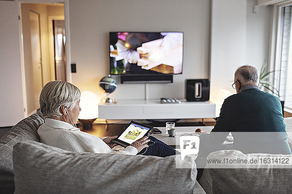 Ältere Frau benutzt digitales Tablet  während ihr Partner auf dem Sofa im Wohnzimmer sitzt