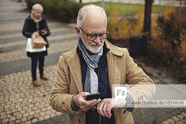 Älterer Mann mit Smartphone  der die Zeit überprüft  während sein Partner auf dem Fußweg hinter ihm steht