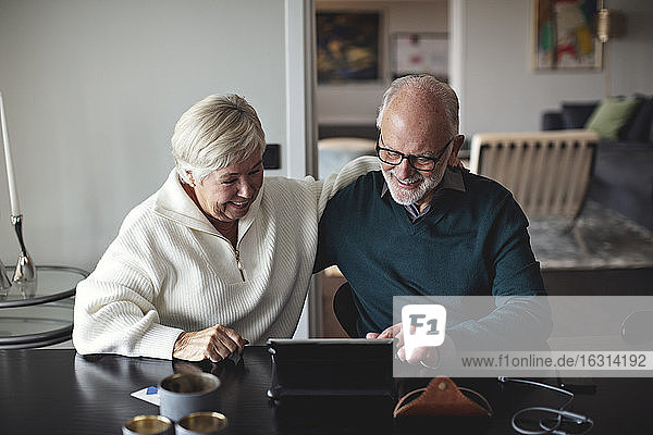 Lächelndes älteres Ehepaar benutzt digitales Tablett  während es am Esstisch im Wohnzimmer sitzt