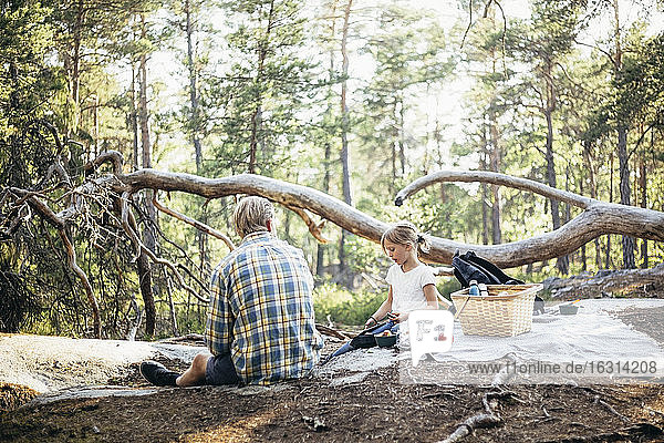 Daughter sitting with father on picnic blanket and using phone in forest